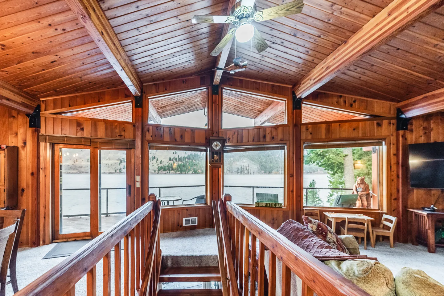 Living room interior featuring wood-paneled walls and ceiling, exposed beams, and large windows looking out at a lake.
