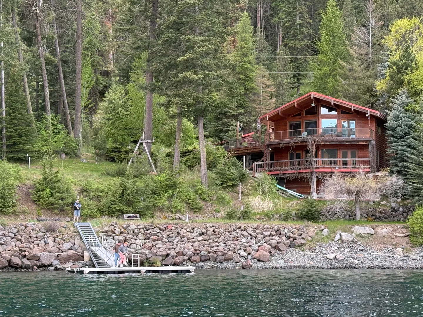 Waterfront view of a multi-story log cabin home set into a forested hillside with a private dock on the water.