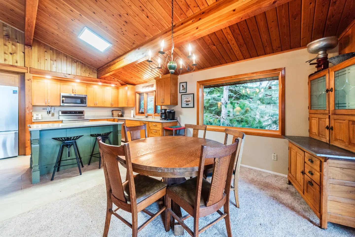Dining area with a round wooden table and chairs, adjacent to a kitchen with wood cabinets and a teal island beneath a vaulted wood ceiling.