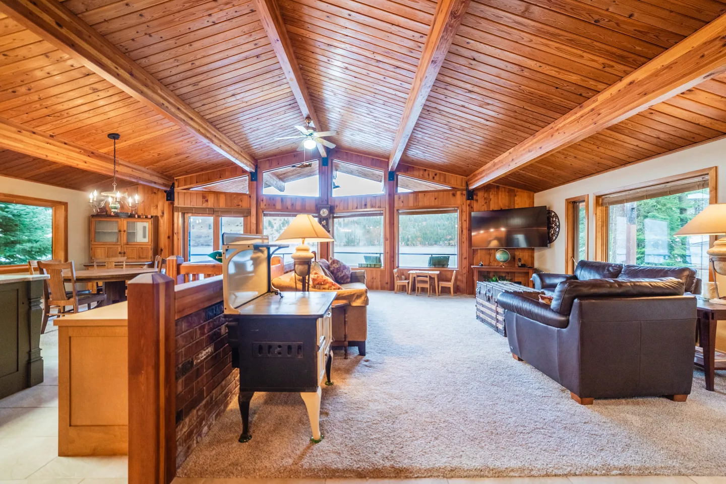 Living room with vaulted wood-paneled ceiling, exposed beams, carpeted floor, leather armchairs, and a wall of windows looking out to the water.