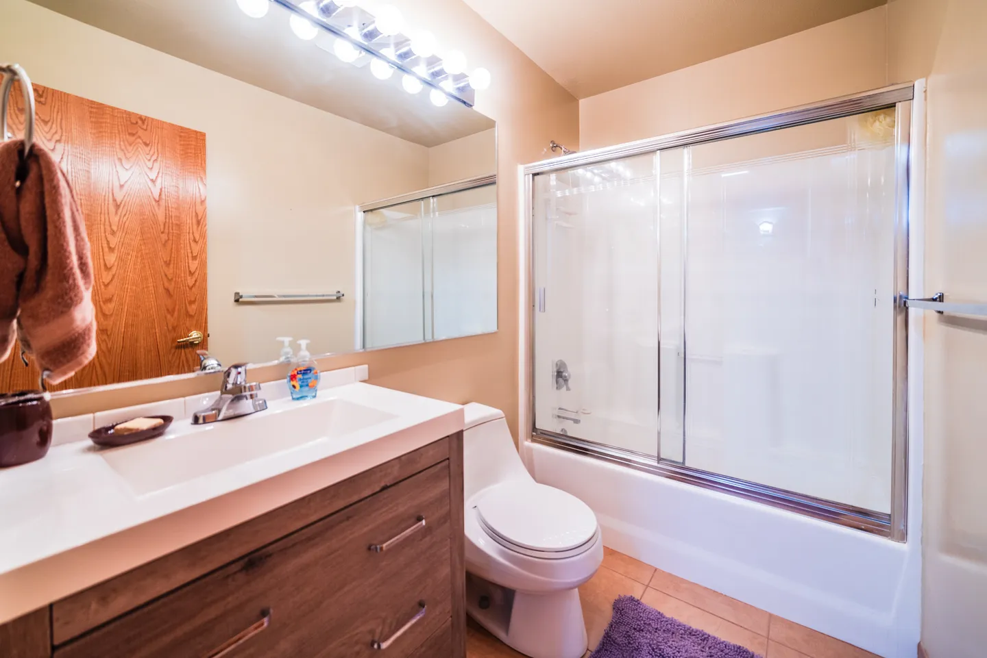 Bathroom featuring a vanity with a white countertop and wood-grain cabinet, a toilet, a large mirror with vanity lighting, and a bathtub with sliding glass doors.