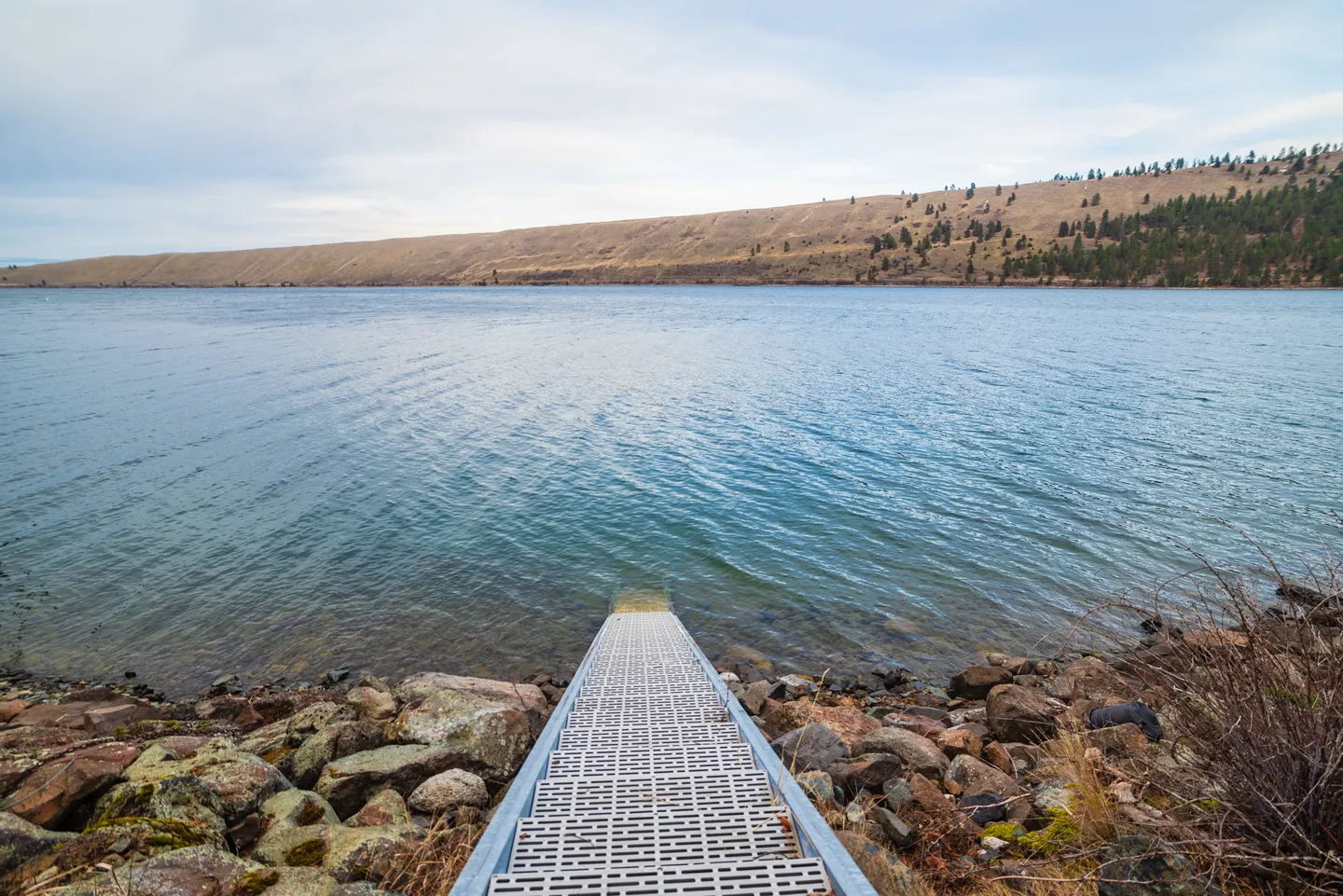A metal ramp leads down rocky shoreline into wide blue water with a dry, rolling hill on the opposite shore.