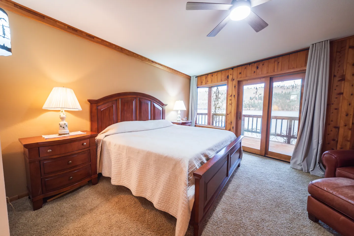 Bedroom with a large wooden bed, matching nightstands with lamps, carpet flooring, and glass doors opening to a balcony with water views.