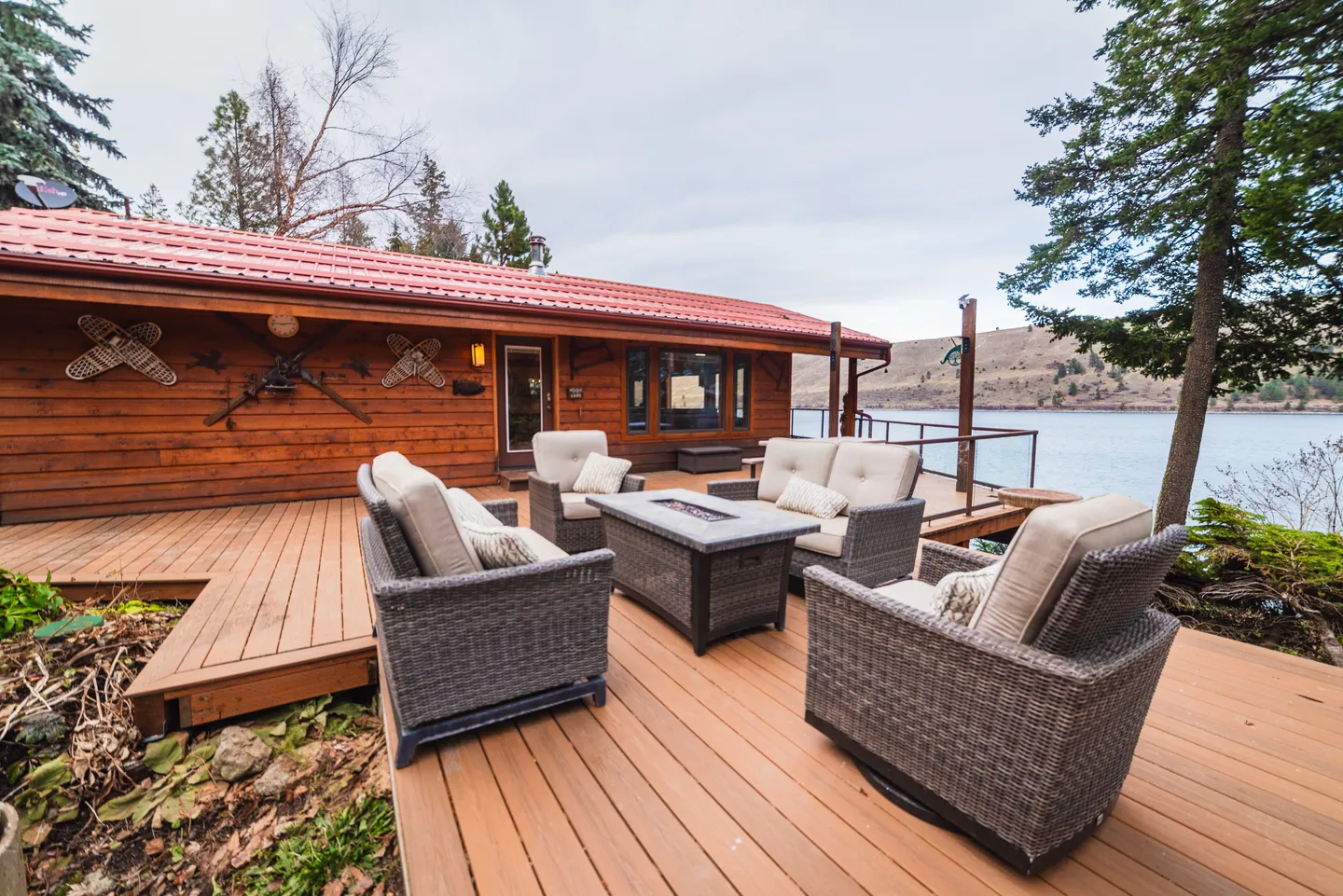 Outdoor wooden deck with wicker seating and a fire table overlooking a lake in front of a log cabin.