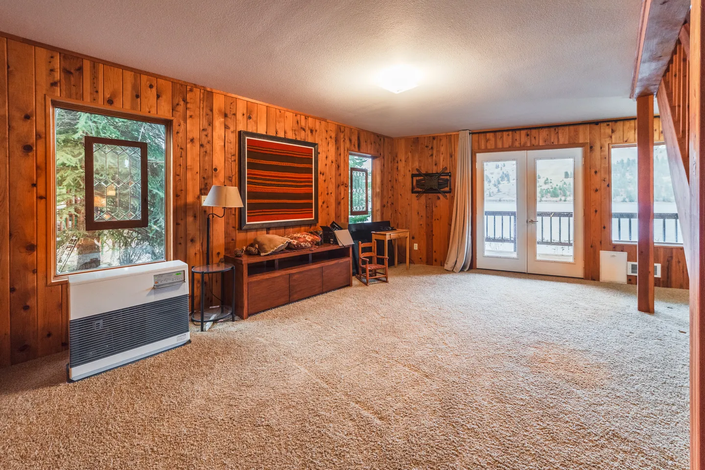 Open room with carpeted floor, wood-paneled walls, low wooden cabinet, floor heater, and glass double doors opening to an outdoor deck.