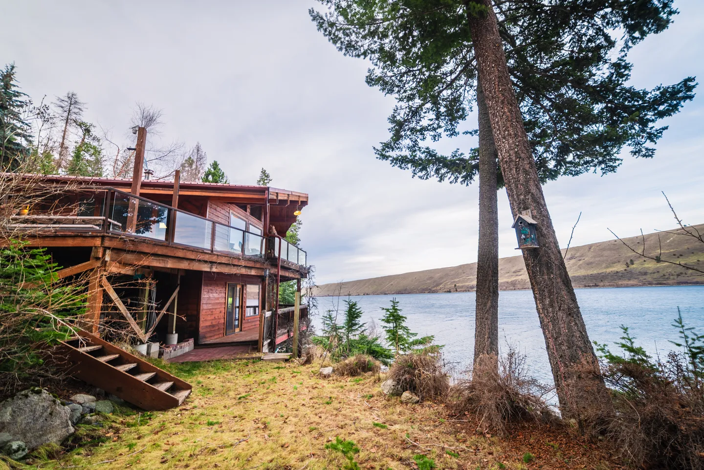 Exterior view of a multi-level wood cabin with an upper glass-railing deck overlooking a lake and distant hills.