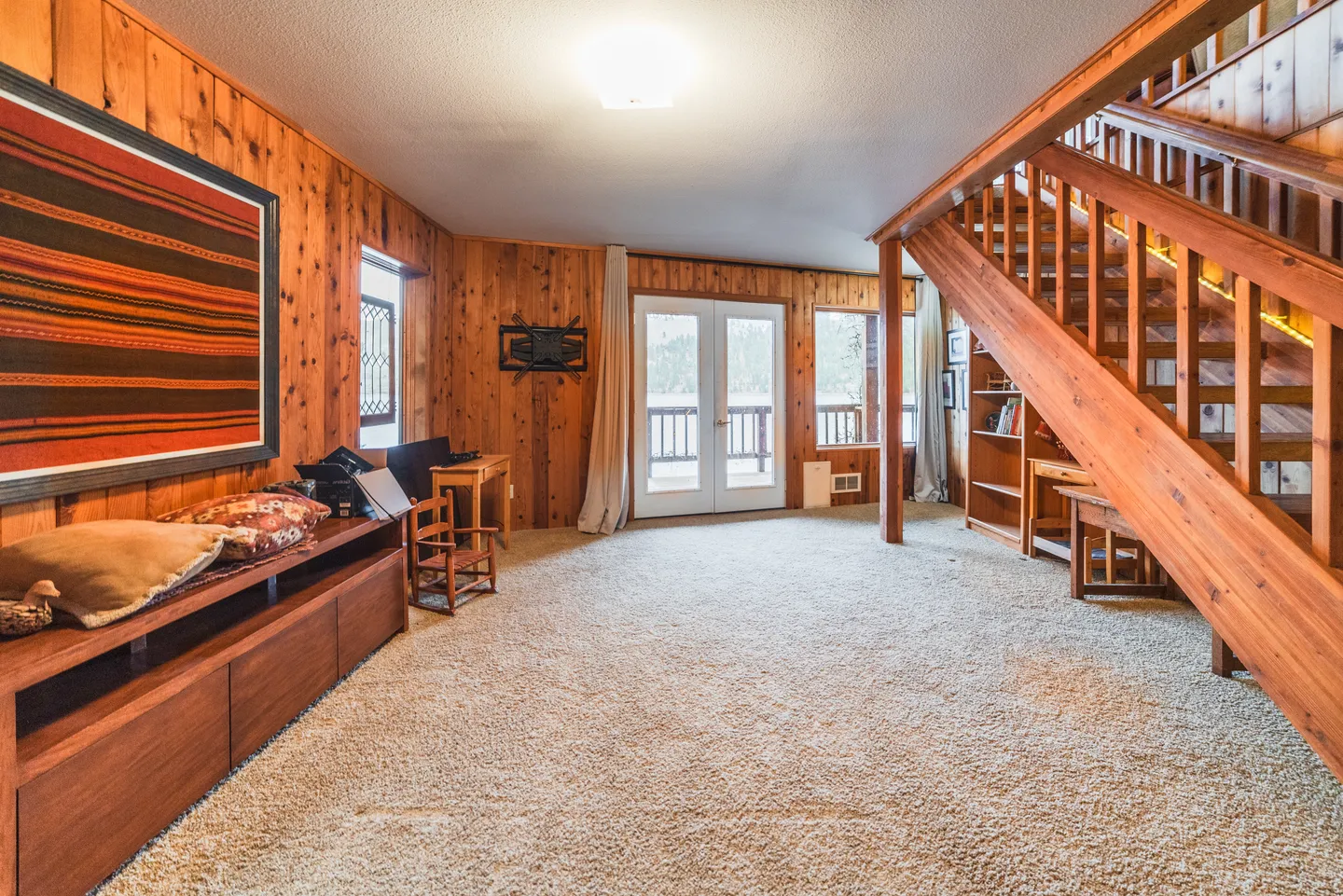 Carpeted living area with wood paneled walls, glass double doors to a deck, and an open wooden staircase.
