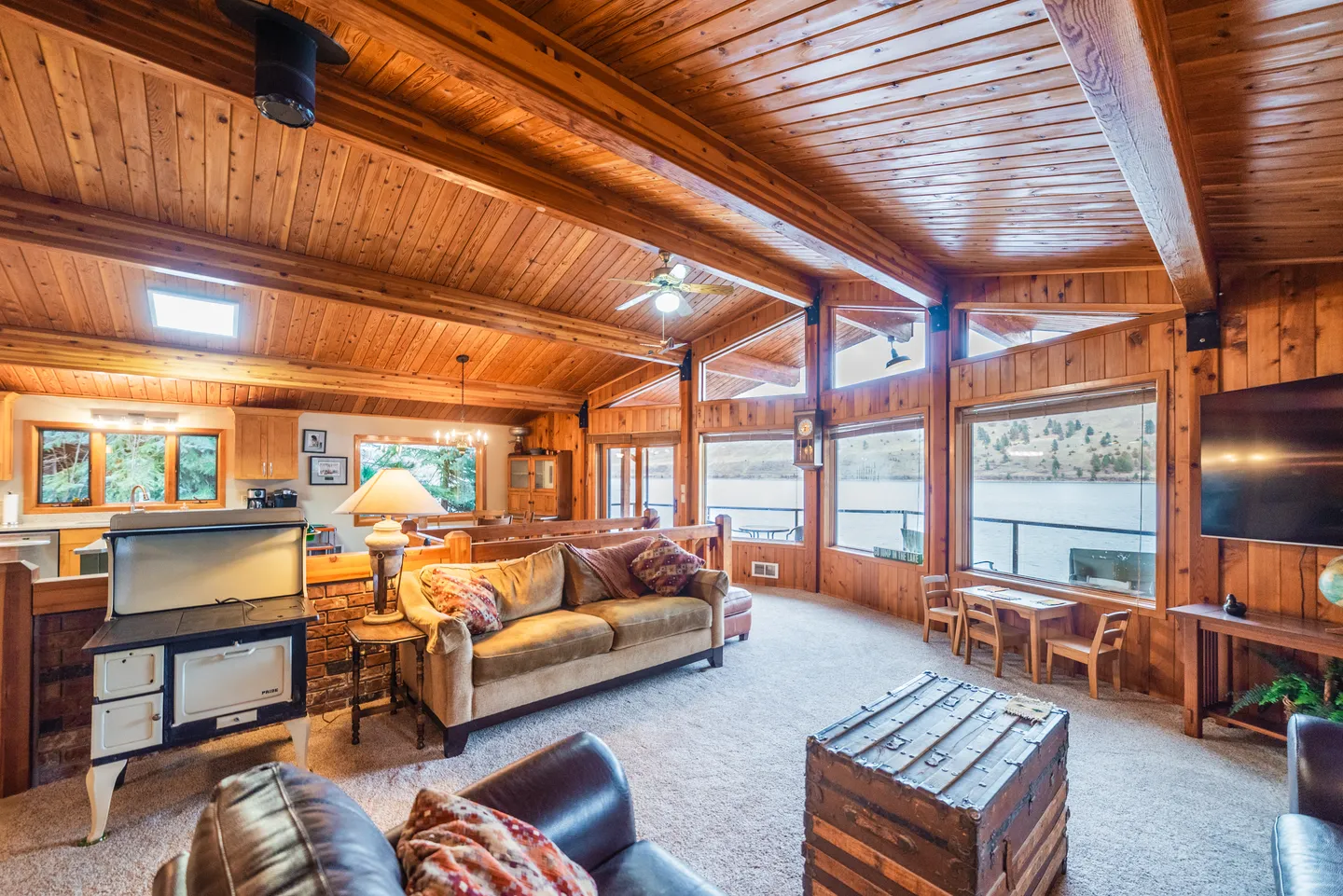 Living room with wood-paneled vaulted ceilings, exposed beams, comfortable seating, and large windows overlooking water.