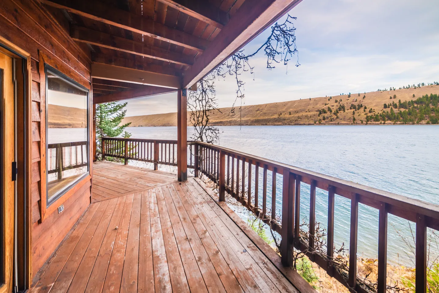 A covered wooden deck with a wooden railing overlooks a wide river and hills under a partly cloudy sky.