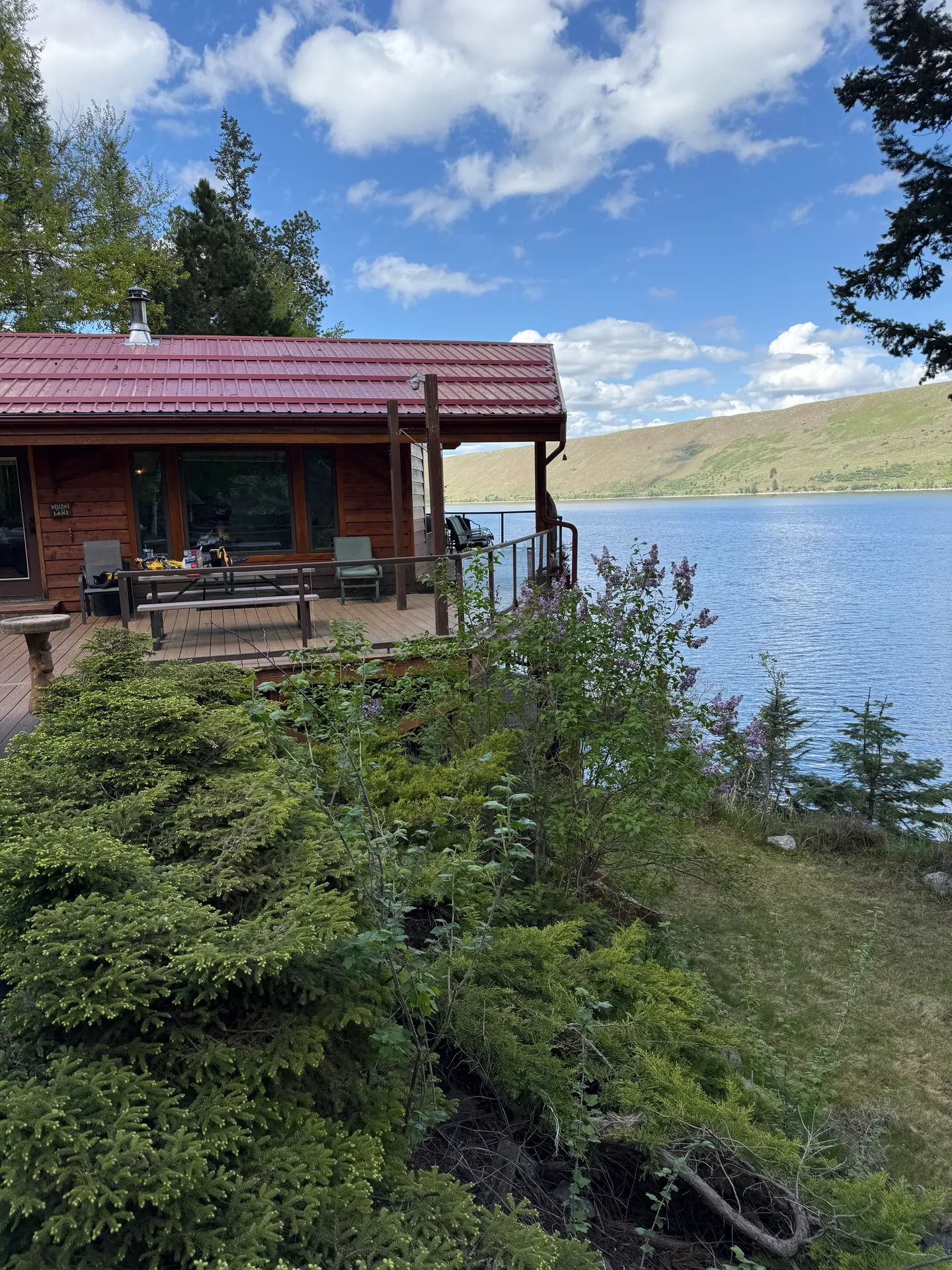 Exterior view of a log cabin with a red metal roof and wraparound deck overlooking a large blue lake and rolling hills.