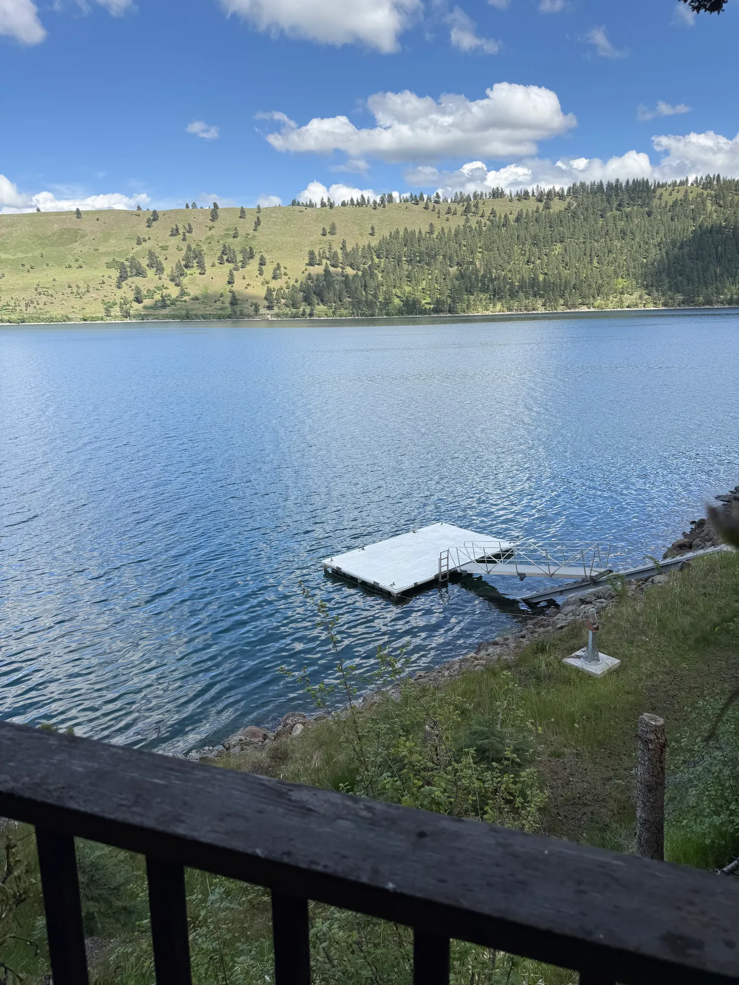 Elevated view from a wooden deck railing looking down at a floating dock on a lake with rolling green hills across the water.