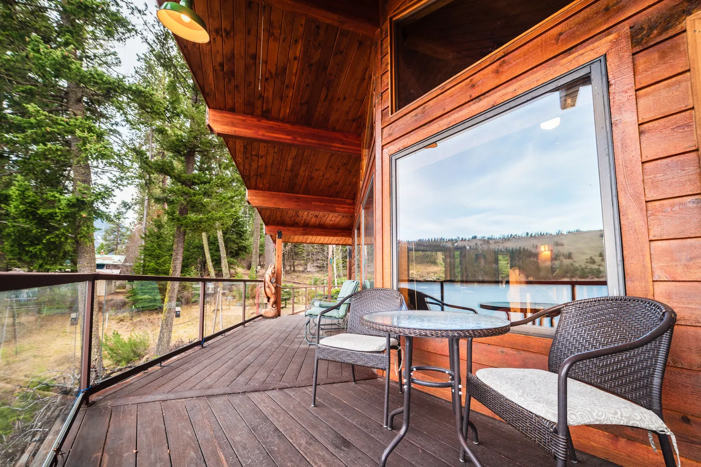 Elevated wooden deck along a log cabin exterior, featuring outdoor chairs, a small table, glass railings, and a view of trees and water.