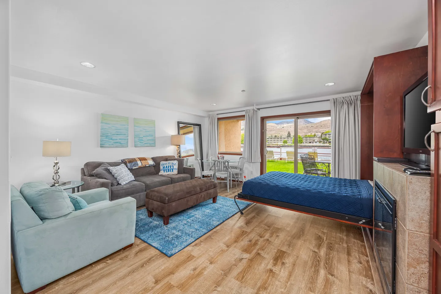 Living area with a sofa, armchair, pull-down Murphy bed, and sliding glass doors opening to a patio and grassy outdoor view.