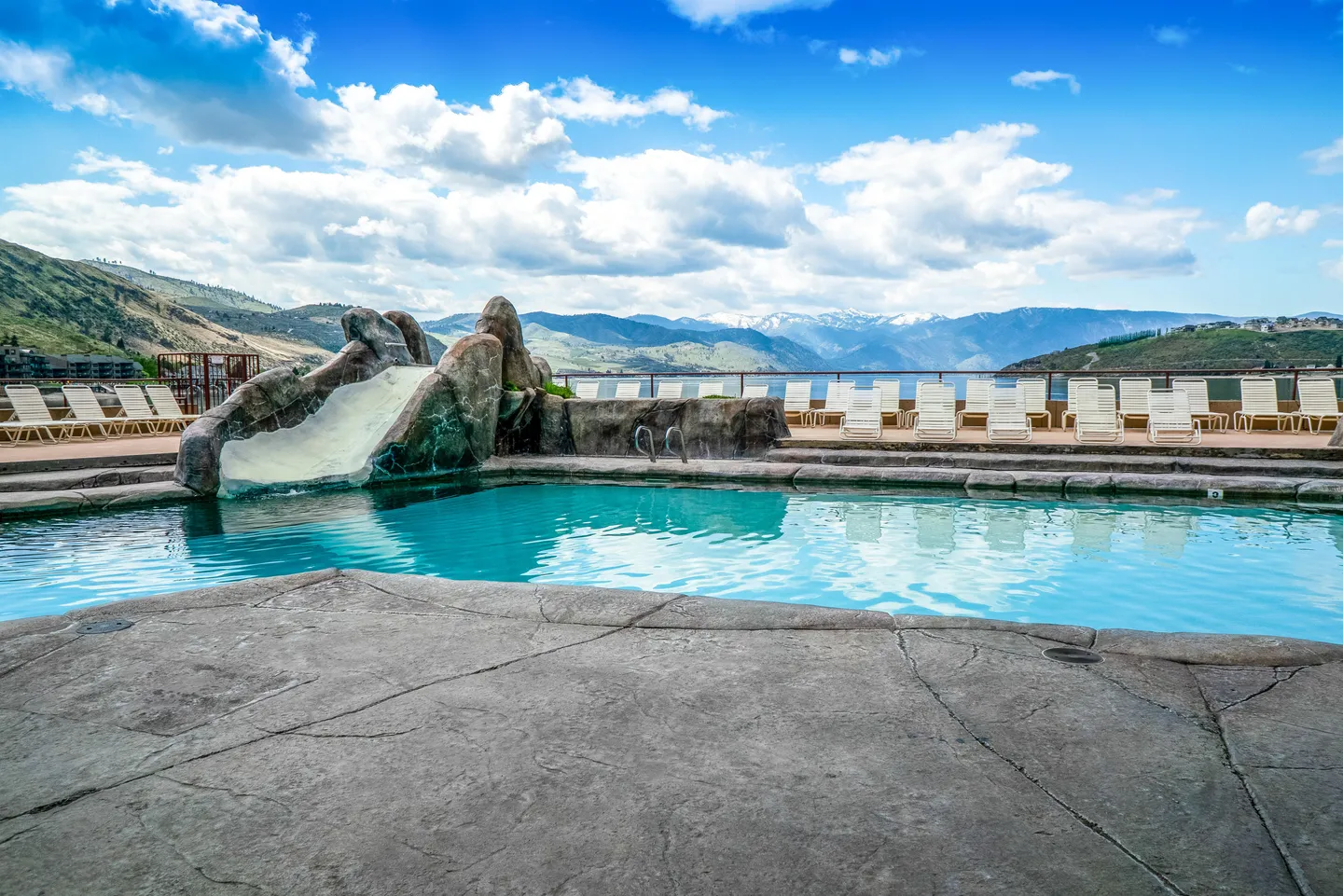 Outdoor swimming pool with a rock water slide, patio lounge chairs, and mountain views under a blue sky with clouds.