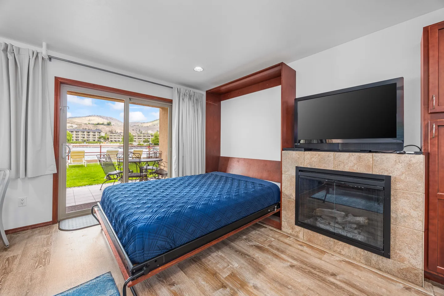 A bedroom area featuring a fold-down Murphy bed with a blue quilt, a tiled fireplace with a TV above it, and sliding glass doors opening to an outdoor patio with mountain views.