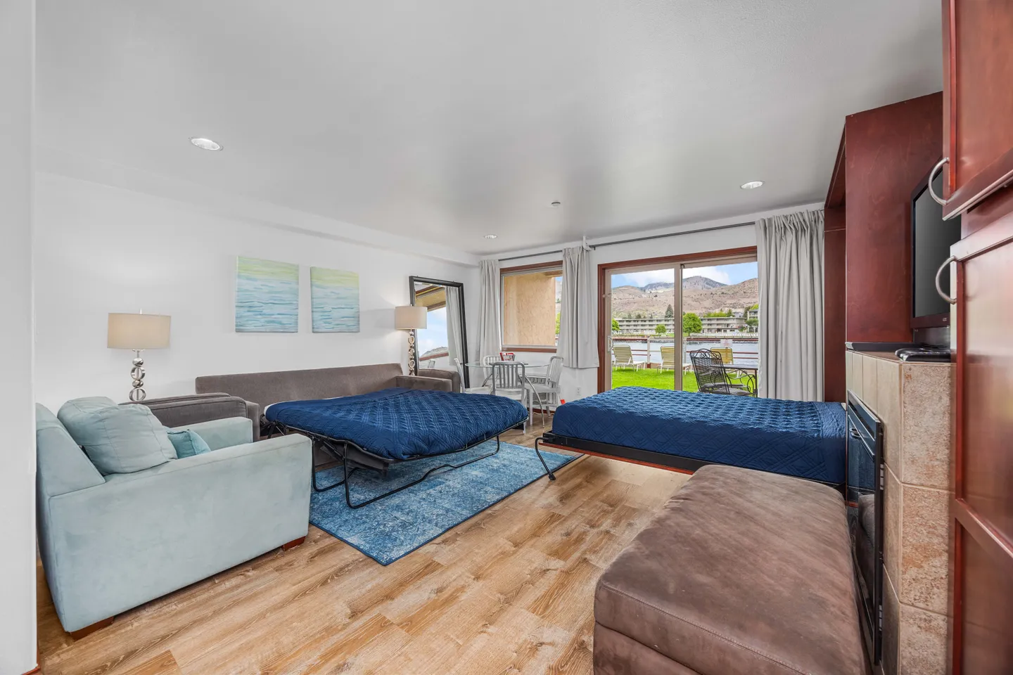 Living area with two pull-out beds extended with blue covers, an armchair, fireplace, and sliding glass doors leading to a patio.