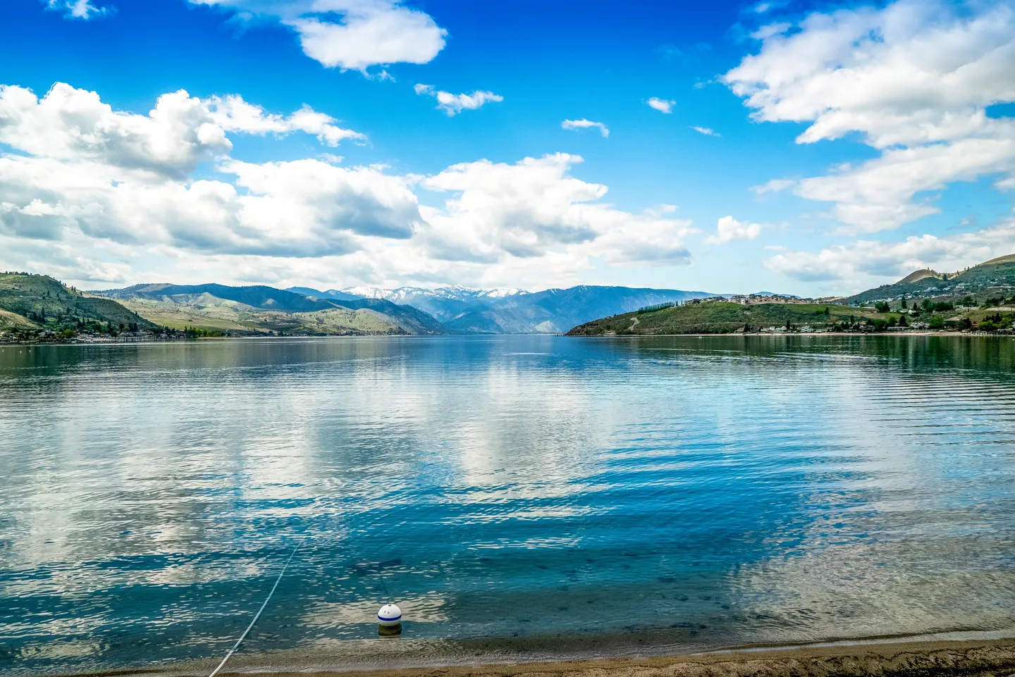 Wide view of a calm blue lake reflecting puffy white clouds, framed by distant mountains and rolling hills under a bright blue sky.