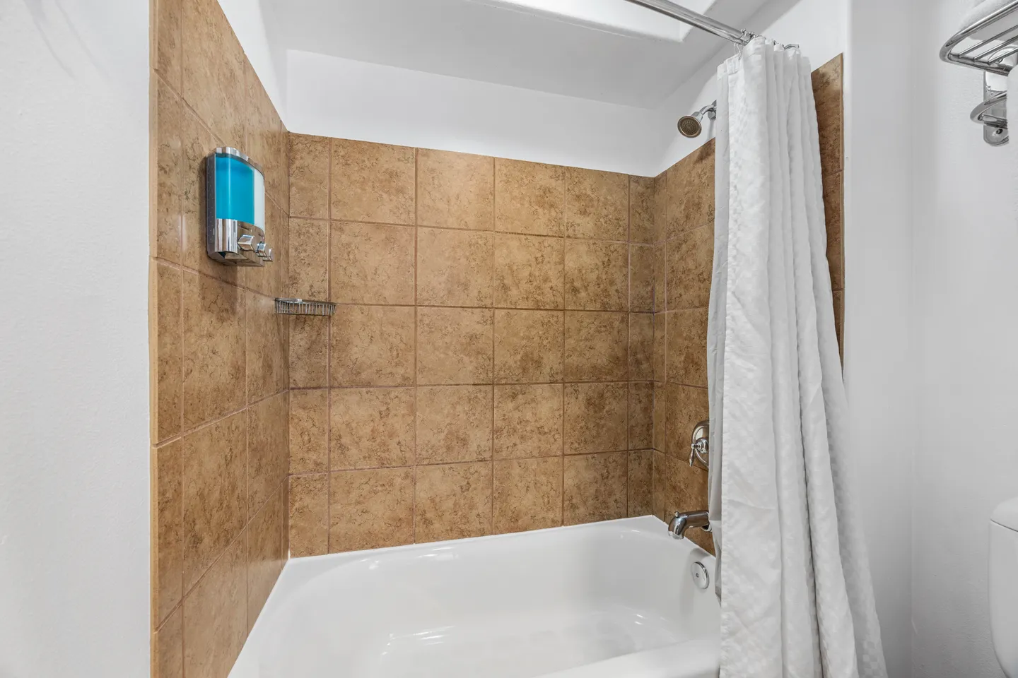 Bathroom shower and bathtub combo surrounded by brown tiles with a white shower curtain and soap dispenser.