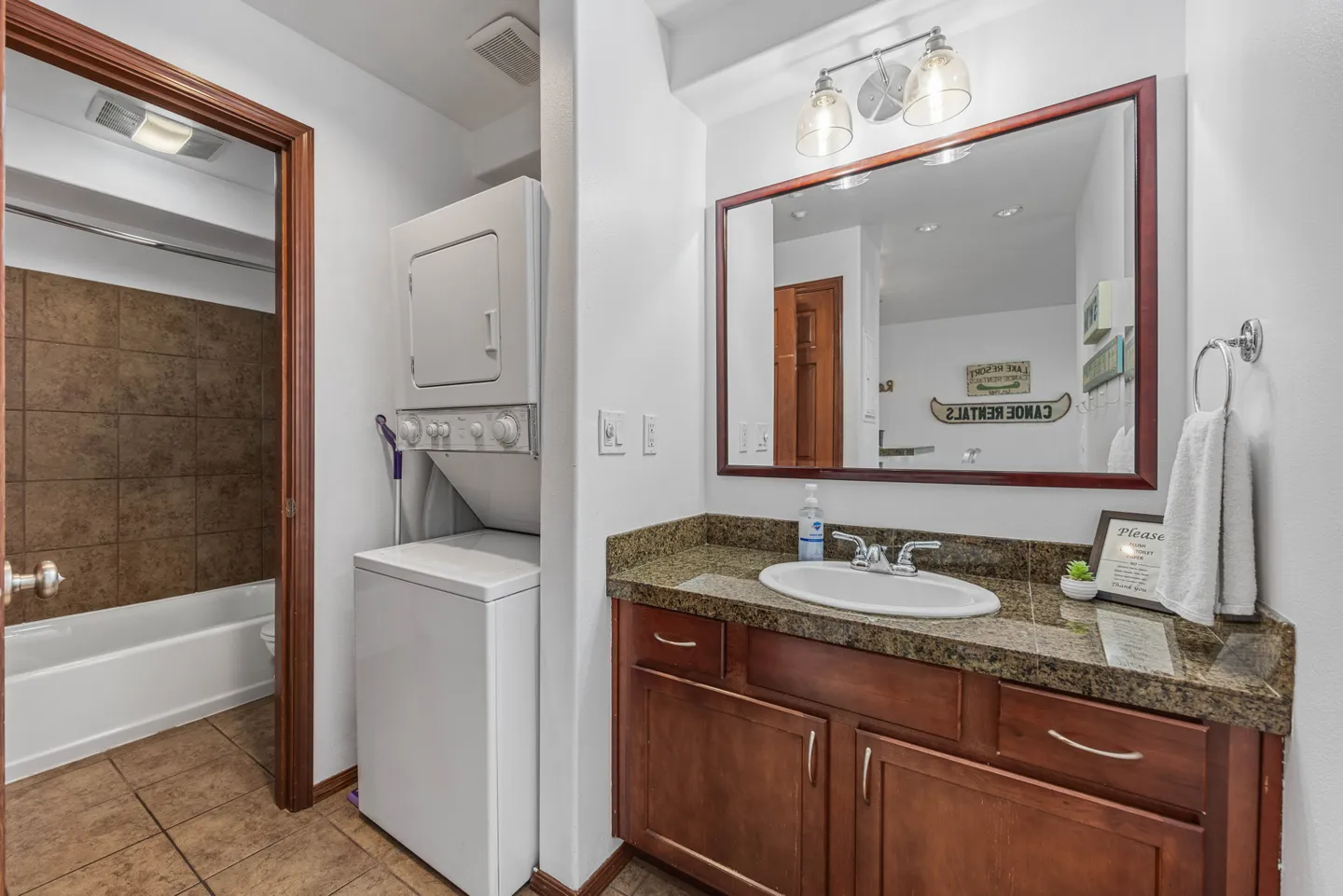 Bathroom with a wood vanity, granite countertop, large framed mirror, stacked washer and dryer, and an open doorway leading to a tiled bathtub area.