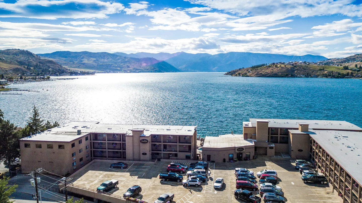 Aerial view of a lakeside multi-story building complex with a parking lot overlooking water and distant mountains.