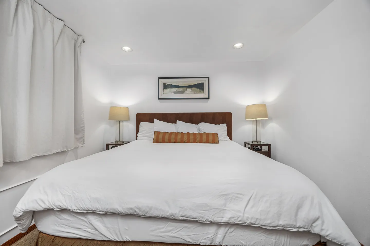 Bedroom with a large bed dressed in white linens, flanked by nightstands with lamps under recessed lighting.