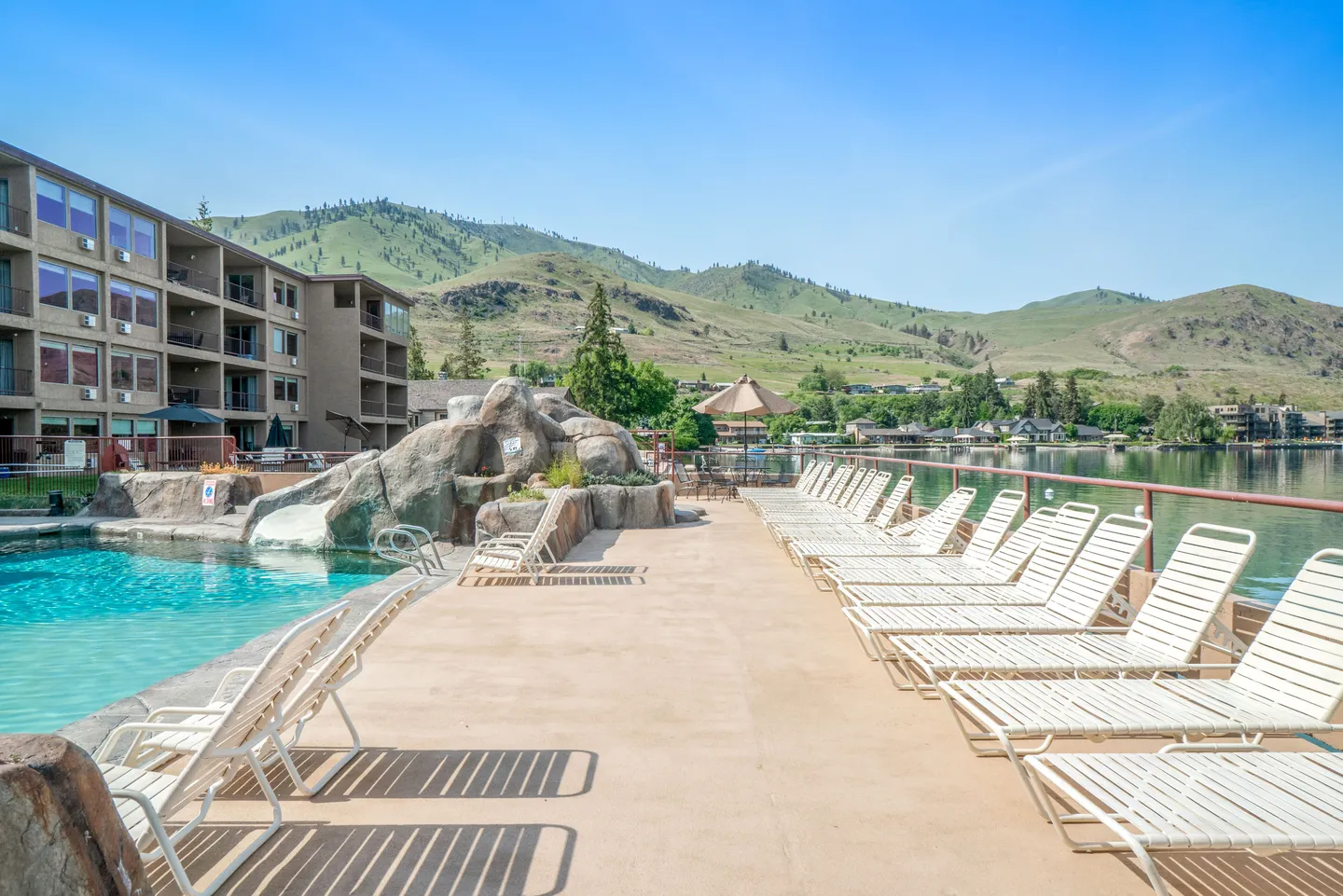Outdoor swimming pool deck with lounge chairs beside a lake, adjacent to a multistory resort building with mountain views.