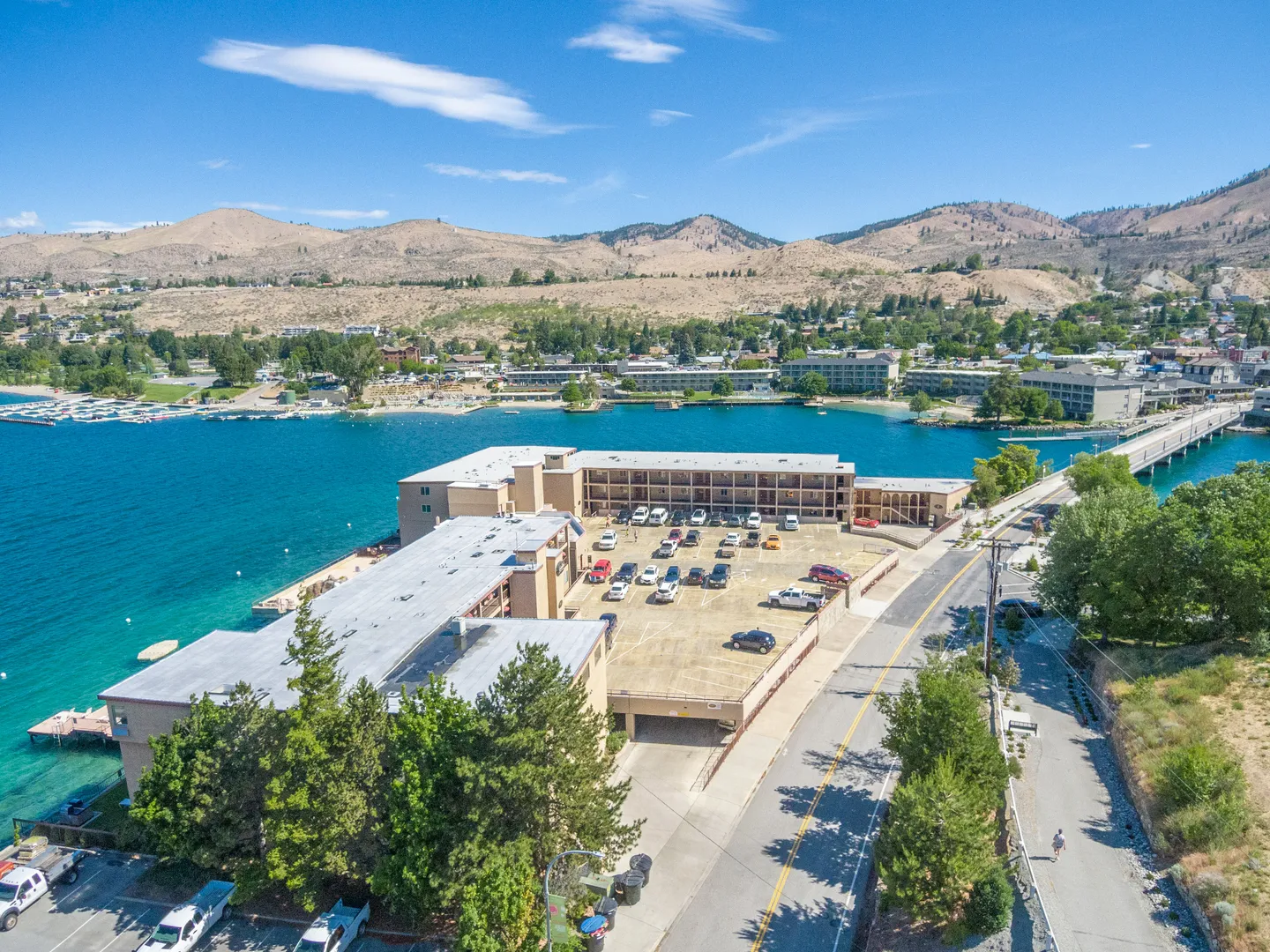 Aerial view of a multi-story waterfront resort building with a large parking lot on a bright blue lake surrounded by hills.