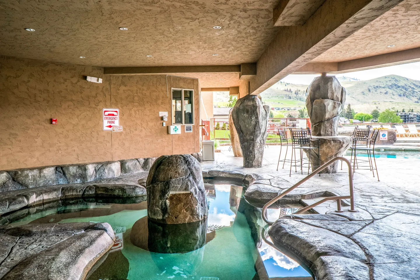 A covered outdoor hot tub featuring faux rock formations and pillars, looking out toward an open-air swimming pool and distant hills.