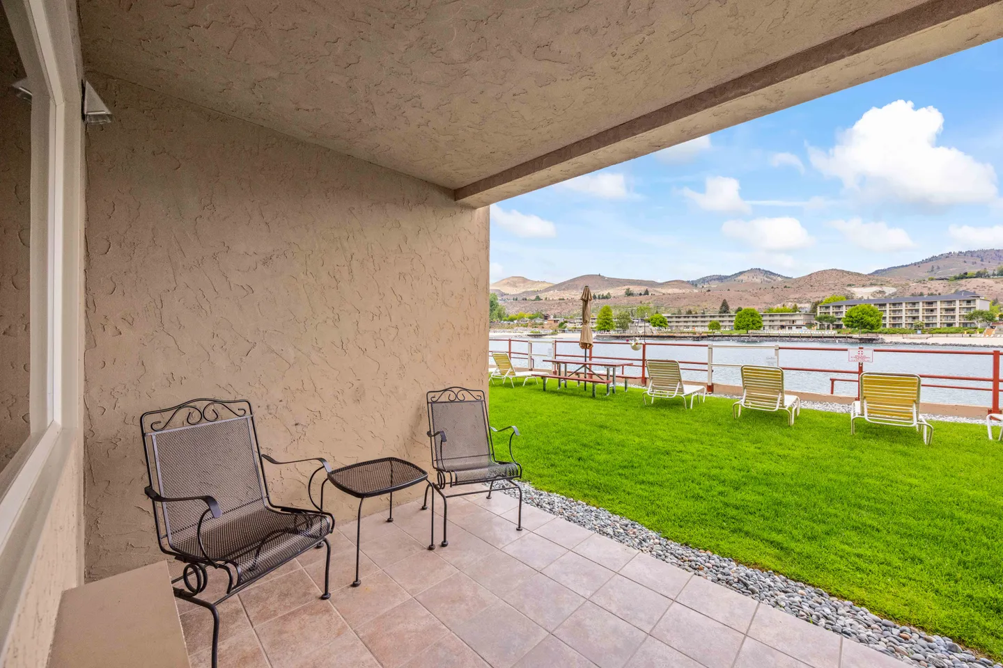 Tiled covered patio with two metal armchairs and a small side table overlooking a lawn, water, and hills.