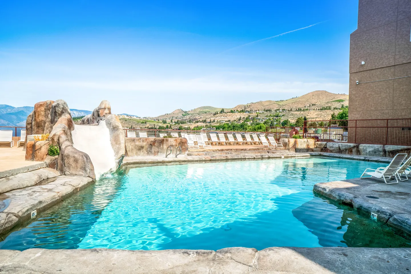 Outdoor swimming pool featuring a rock-style water slide with hills and clear blue sky in the background.