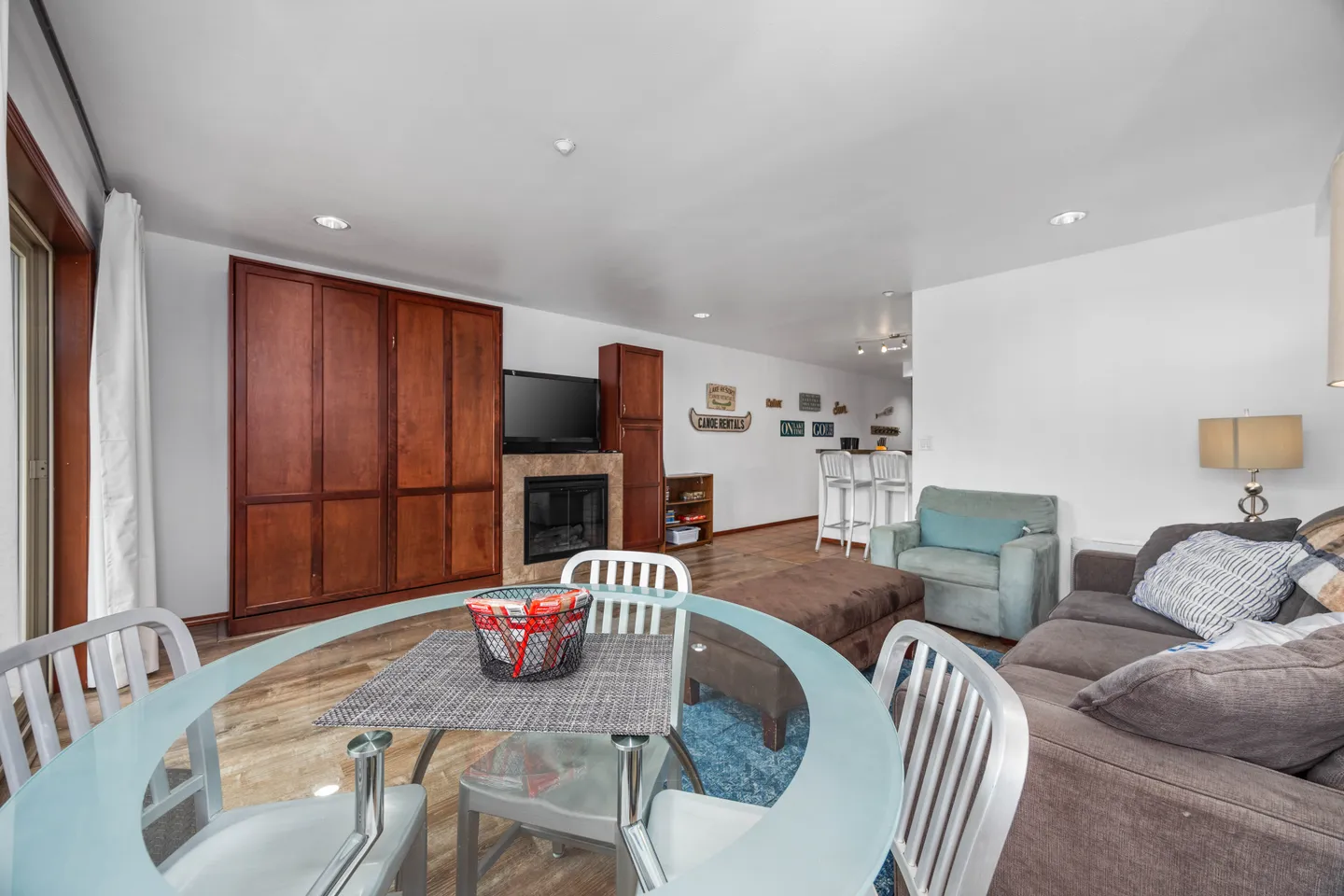 Open-concept living and dining area with a glass dining table in the foreground, sofa, armchair, and built-in wood cabinets surrounding a fireplace.