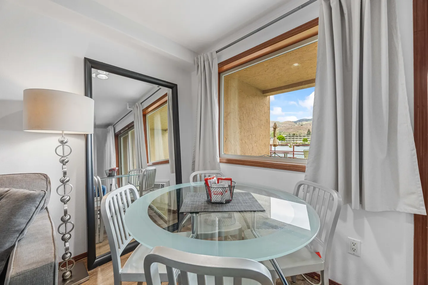 Dining area with a round frosted glass table, metal chairs, a full-length leaning mirror, and a window framing an outdoor view.