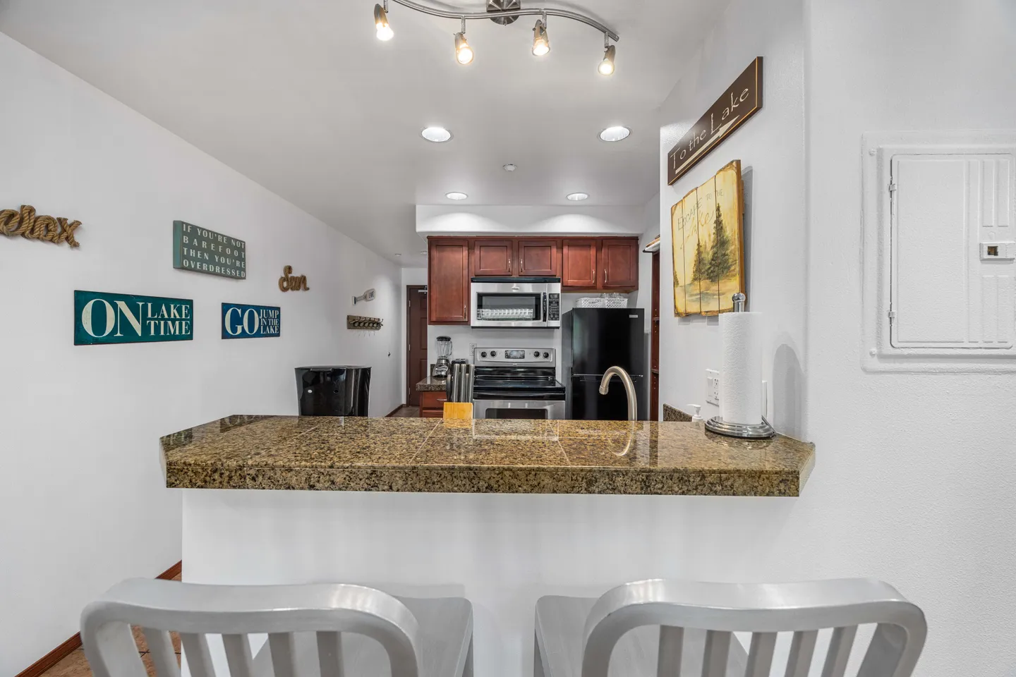 A raised granite-topped breakfast bar with two silver barstools looking into a kitchen with stainless steel appliances and wood cabinets.
