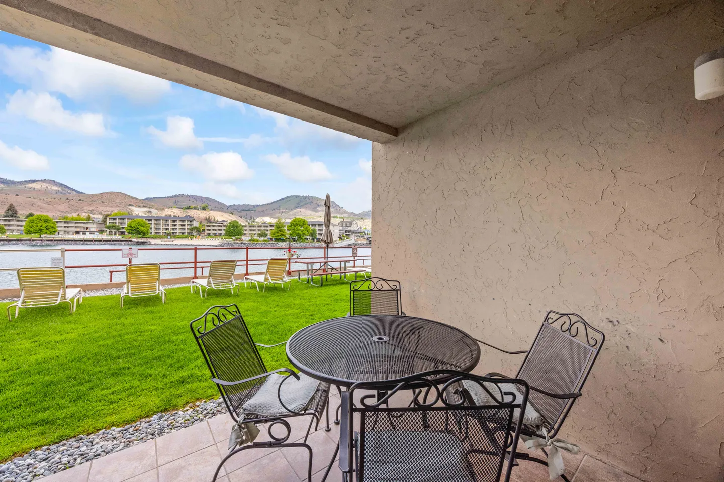 Covered patio with a metal dining set looking out to a grassy lawn, lounge chairs, a body of water, and hills.