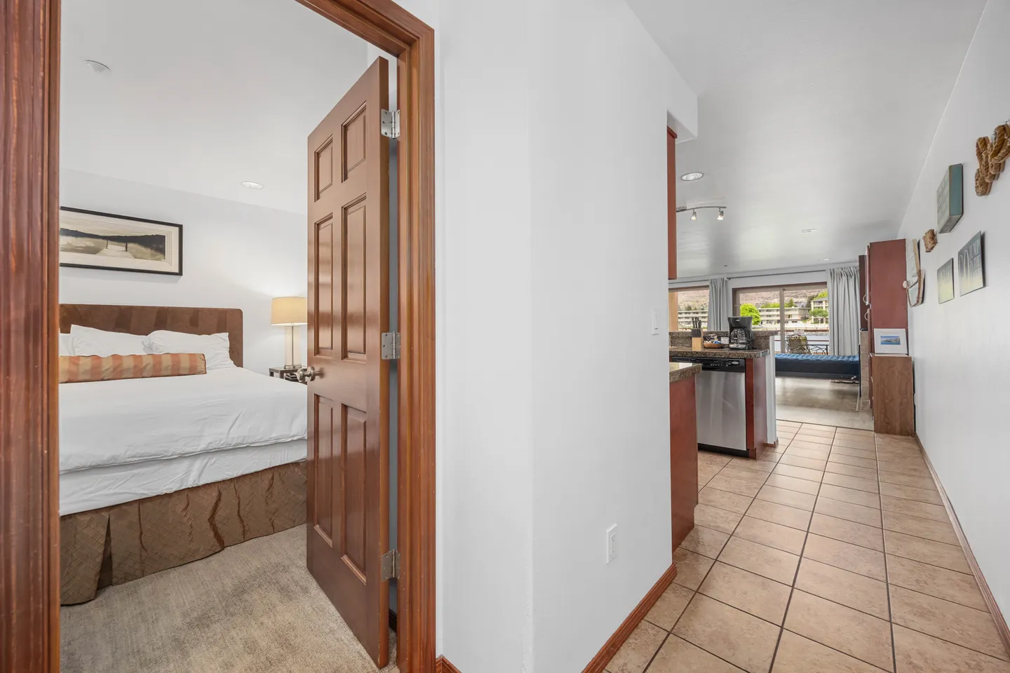 Tiled hallway looking toward a kitchen and living area, with an open doorway revealing a carpeted bedroom with a bed.