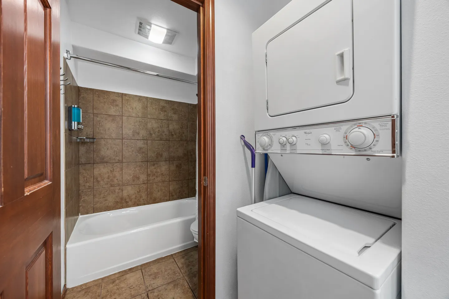 A stacked washer and dryer in the foreground adjacent to an open doorway leading to a tiled bathroom with a tub.