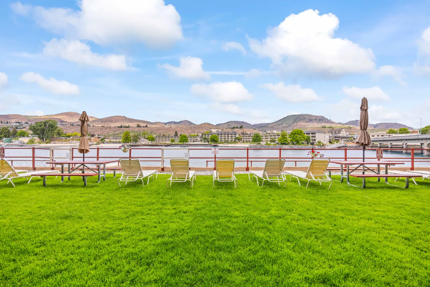 Lawn area with lounge chairs and picnic tables overlooking a lake and surrounding hills under a blue sky.
