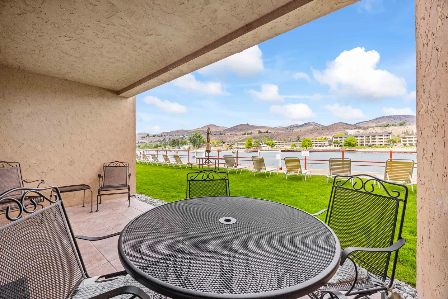 Covered patio with wrought-iron outdoor table and chairs overlooking a green lawn, waterfront, and mountains.