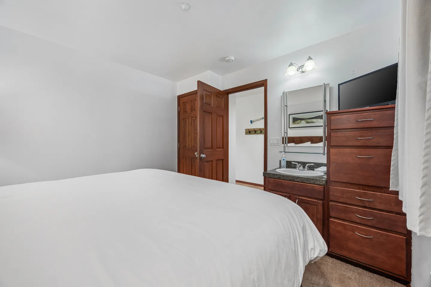 Bedroom featuring a bed with white bedding, a wooden vanity with a sink and mirror, and a matching wooden dresser with a TV.