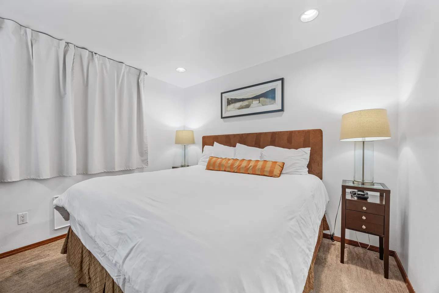 Bedroom with a large bed dressed in white linens, an orange accent pillow, bedside tables with lamps, and recessed ceiling lights.