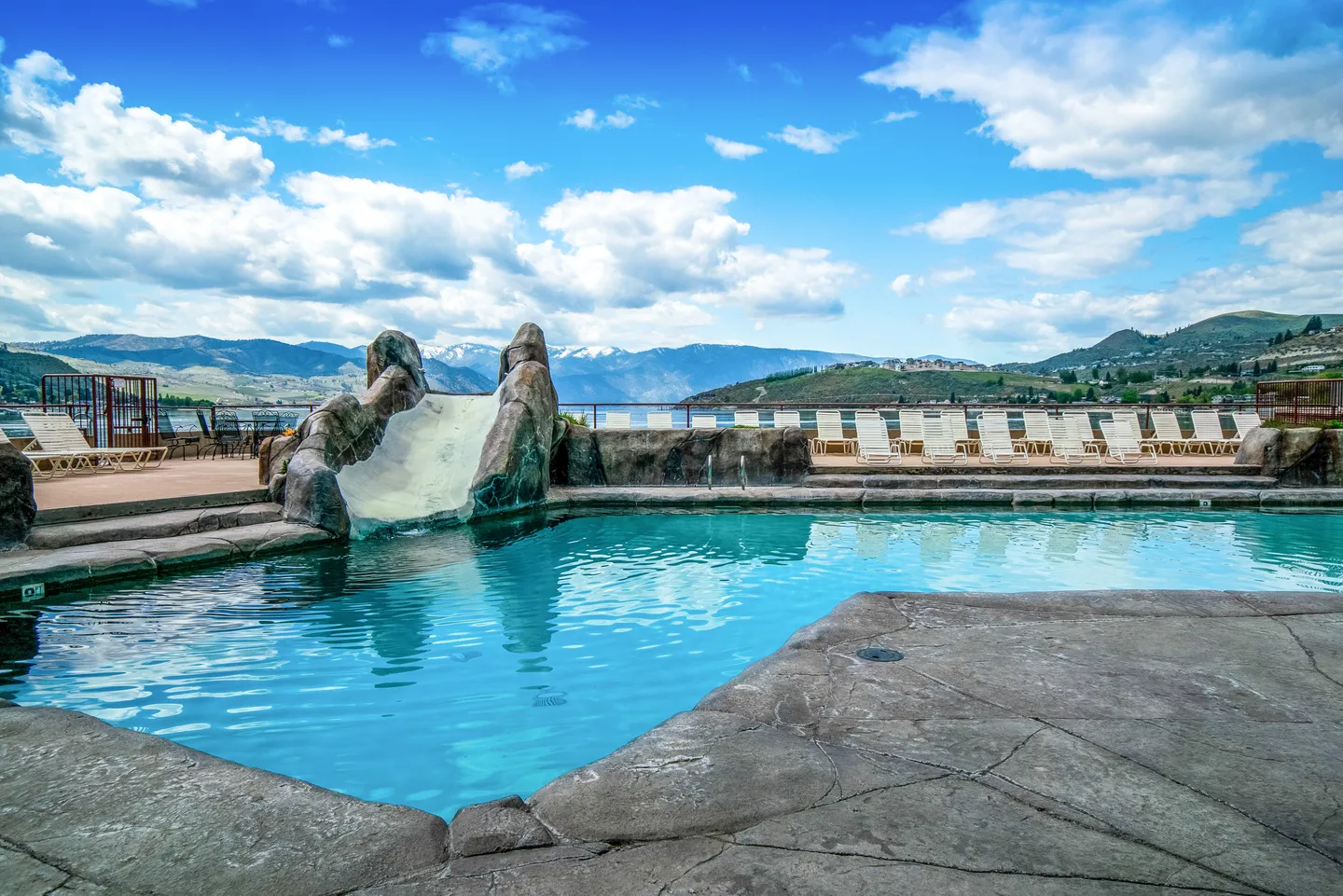 Outdoor swimming pool featuring a rock-style water slide with lounge chairs and a mountain-and-water backdrop under a partly cloudy sky.