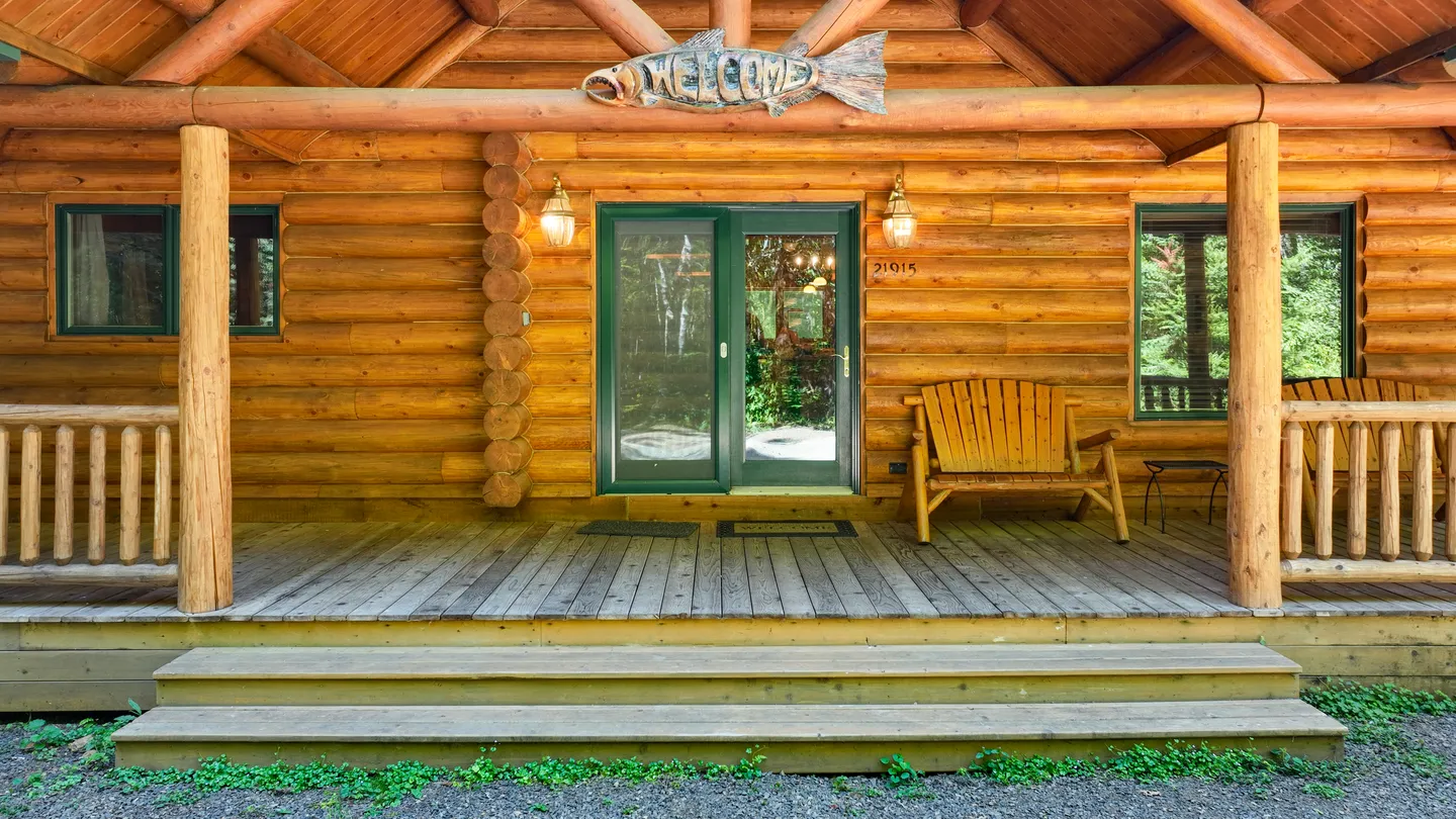 Front porch of a log cabin with wooden steps, a glass entrance door, log railings, and a wooden bench.