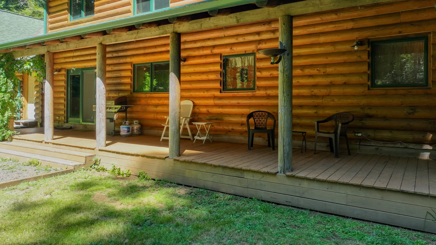 A covered wooden porch along the side of a log cabin with outdoor chairs, a barbecue grill, and a grassy lawn in the foreground.