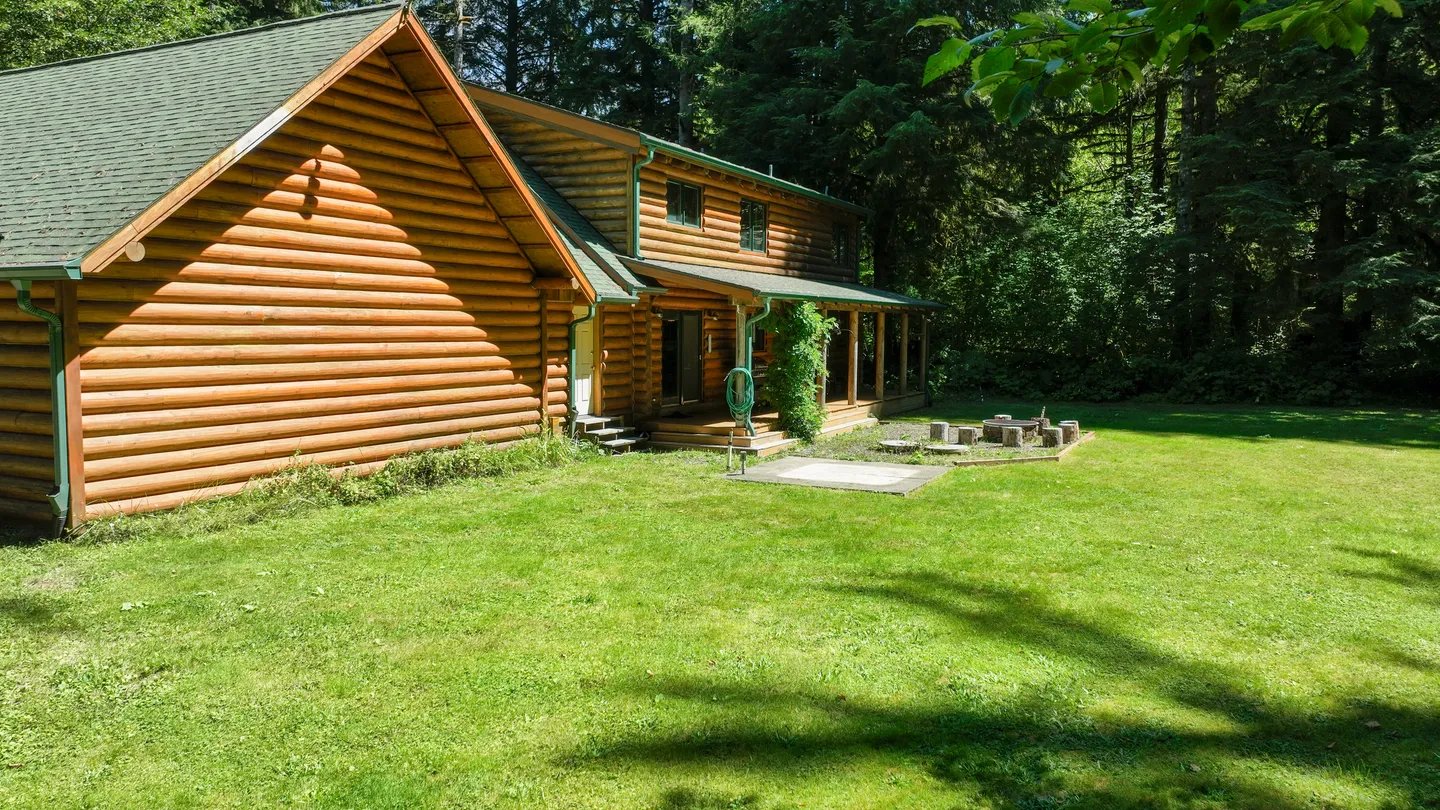 Exterior of a two-story log cabin with a covered back porch and a large grassy yard bordered by dense trees.