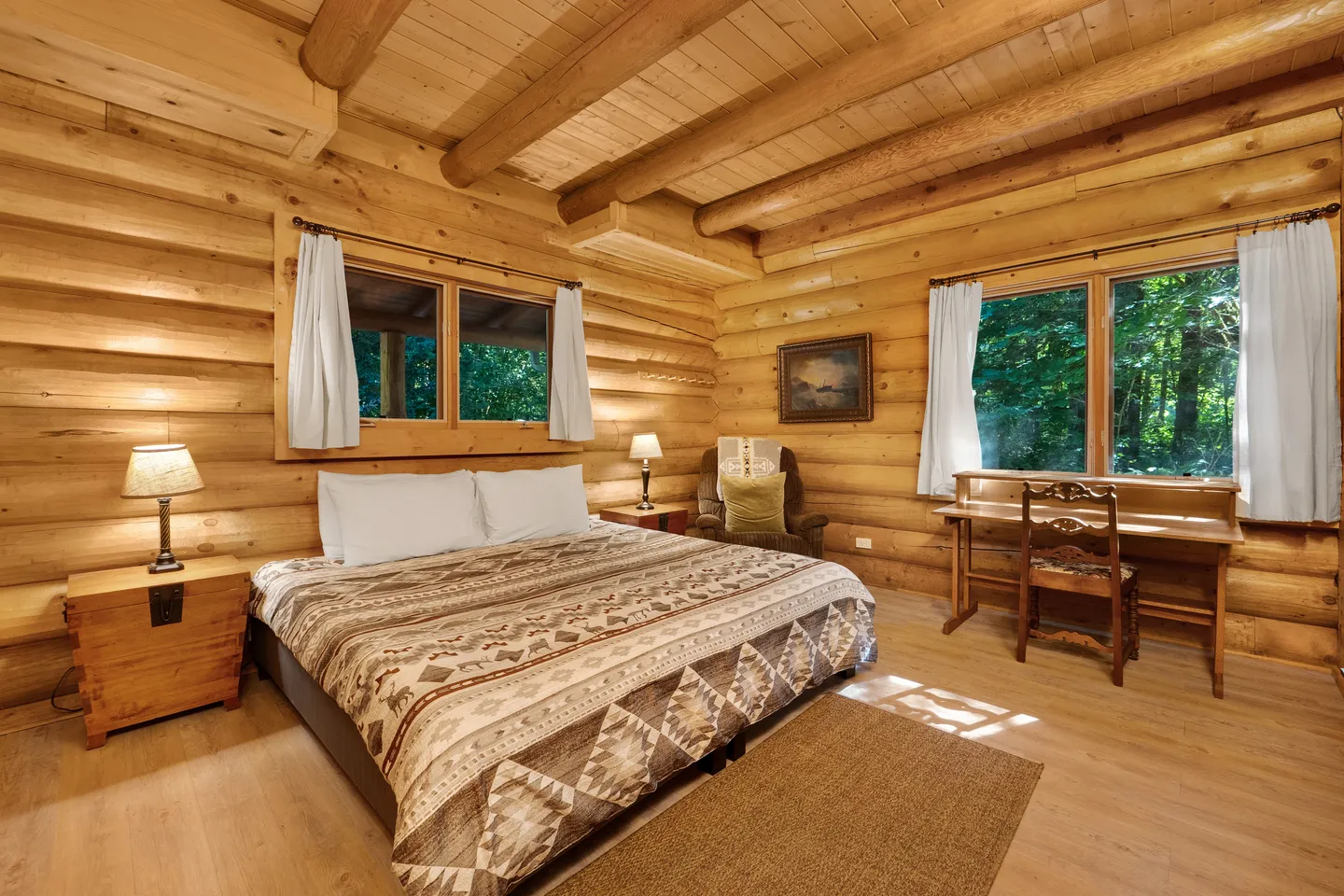 Log cabin bedroom with a patterned bedspread, wooden desk, armchair, and log walls.