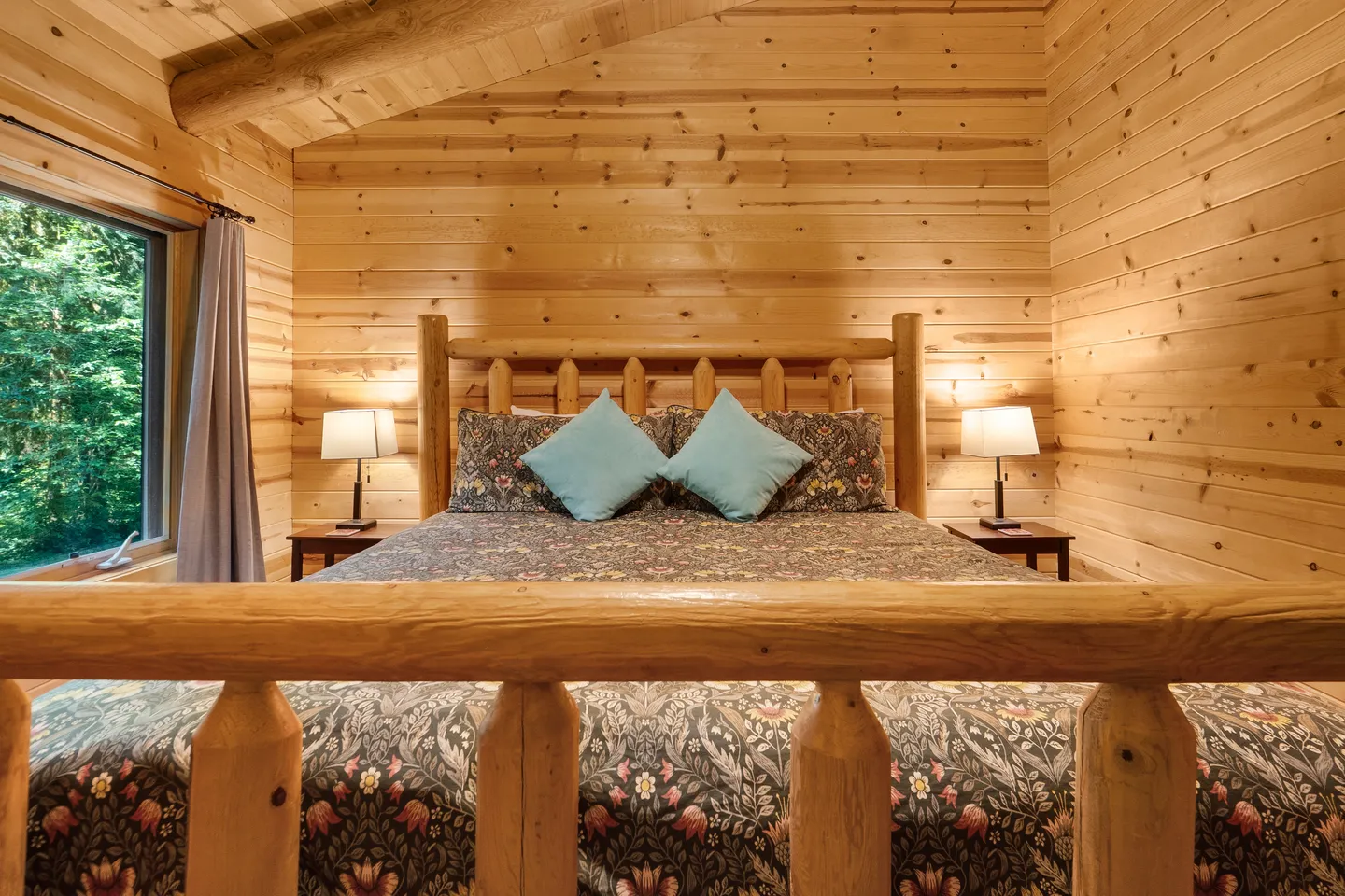 Bedroom with wooden log walls, a wooden log bed frame, nightstands with lamps, and a window showing green trees outside.