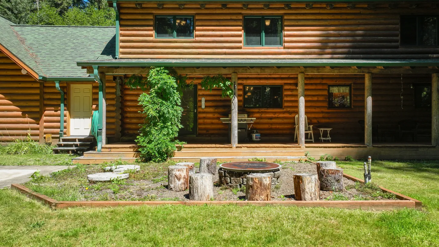Exterior view of a log cabin with a covered front porch and an outdoor fire pit encircled by tree stump seats.