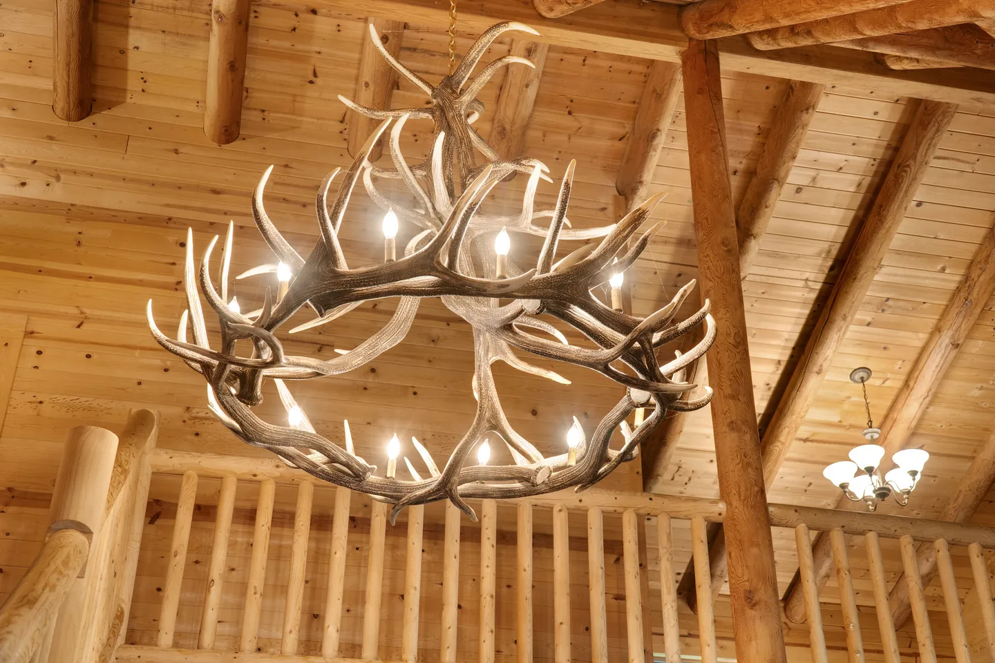 A large lit antler chandelier suspended beneath a natural wood log-beamed ceiling and adjacent to a log railing.