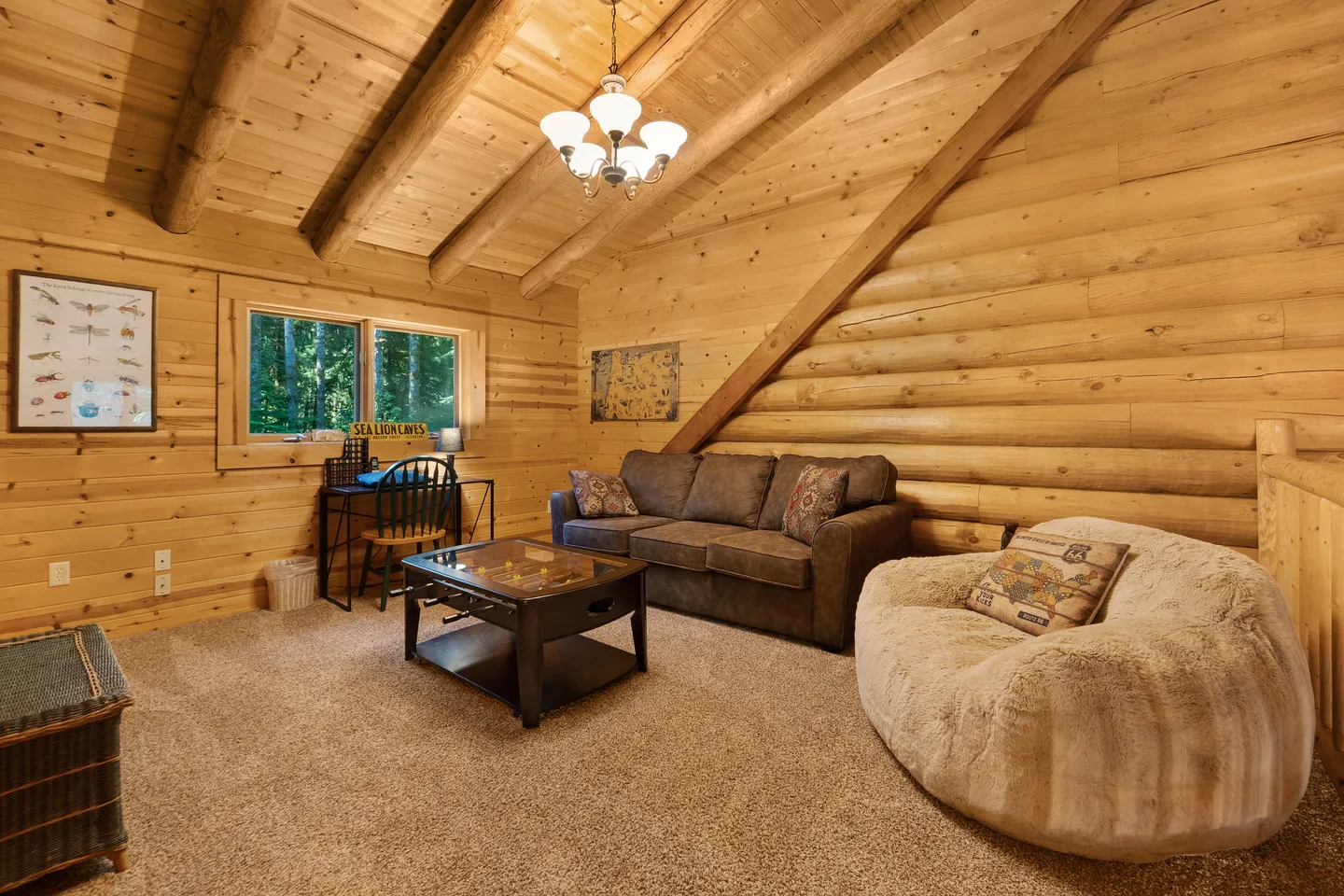 A carpeted log cabin loft living space featuring a brown sofa, oversized plush bean bag chair, small desk, and coffee table beneath a sloped beamed ceiling.