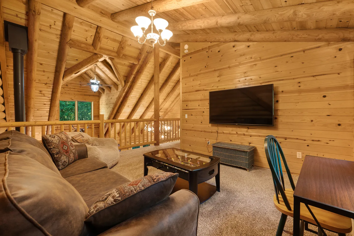 Upstairs loft lounge area with log beam ceilings, carpeted floor, sofa, wall-mounted television, and foosball coffee table.